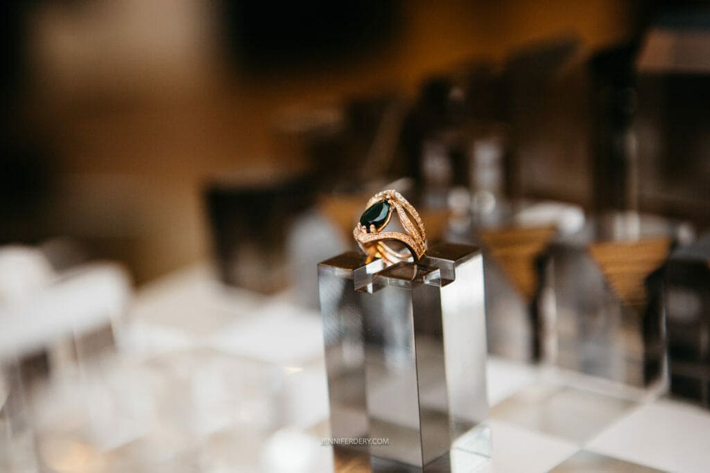 A gold ring with a dark gemstone is displayed on a reflective, metallic stand. The ring has intricate detailing and is surrounded by various geometric and angular objects, creating an elegant and modern arrangement.