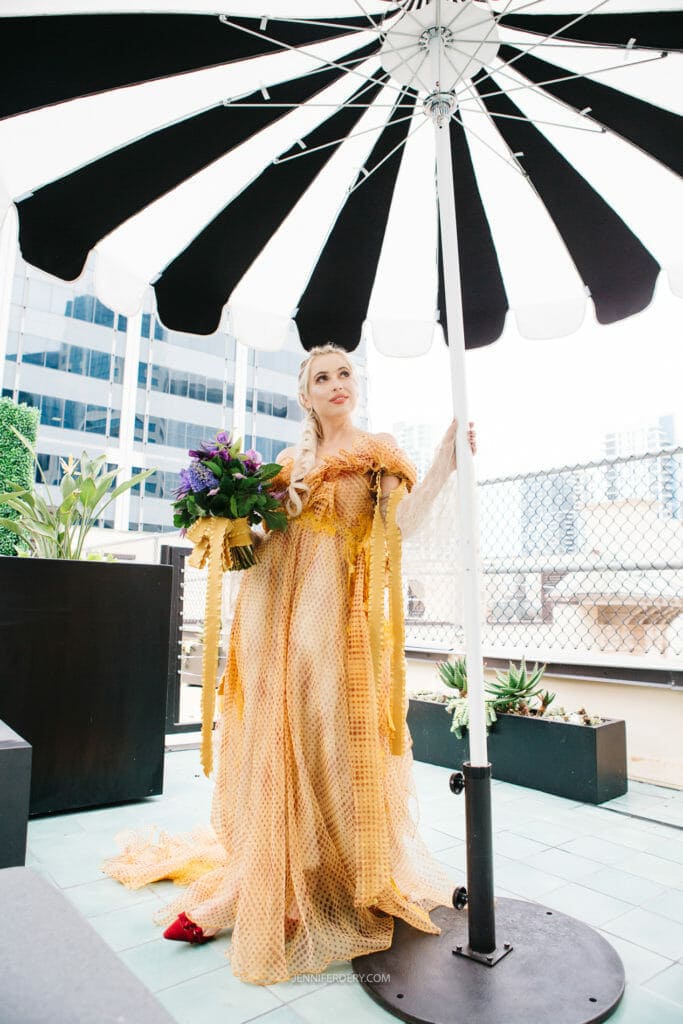 A bride in an elaborate, gold, netted gown stands on a rooftop patio under a black-and-white umbrella. She holds a bouquet of flowers and looks upwards. The background features tall buildings and some plants in pots.
