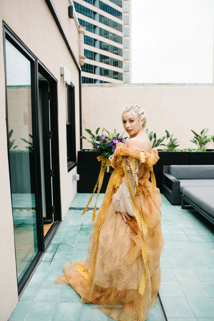 A bride stands on a patio in a flowing, yellow, netted dress with intricate details, holding a bouquet of purple flowers. The individual has light-colored braided hair and looks over their shoulder at the camera. The background features tall buildings and patio furniture.