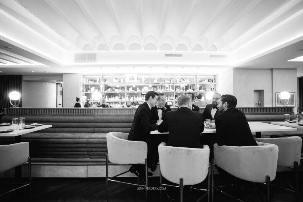 A group of groomsmen in suits are seated around a table in at the historic guild hotel, engaging in conversation. The interior is elegant with a well-lit bar and shelves of bottles in the background. The ceiling has unique geometric designs, and there are unoccupied chairs nearby.