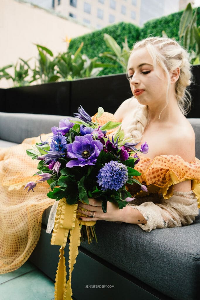 A woman with long blonde hair in a braid is seated on a gray sofa, holding a vibrant flower bouquet with purple and blue blossoms and green leaves. She is wearing an off-shoulder, yellow dress with orange accents. Plant greenery is visible in the background.