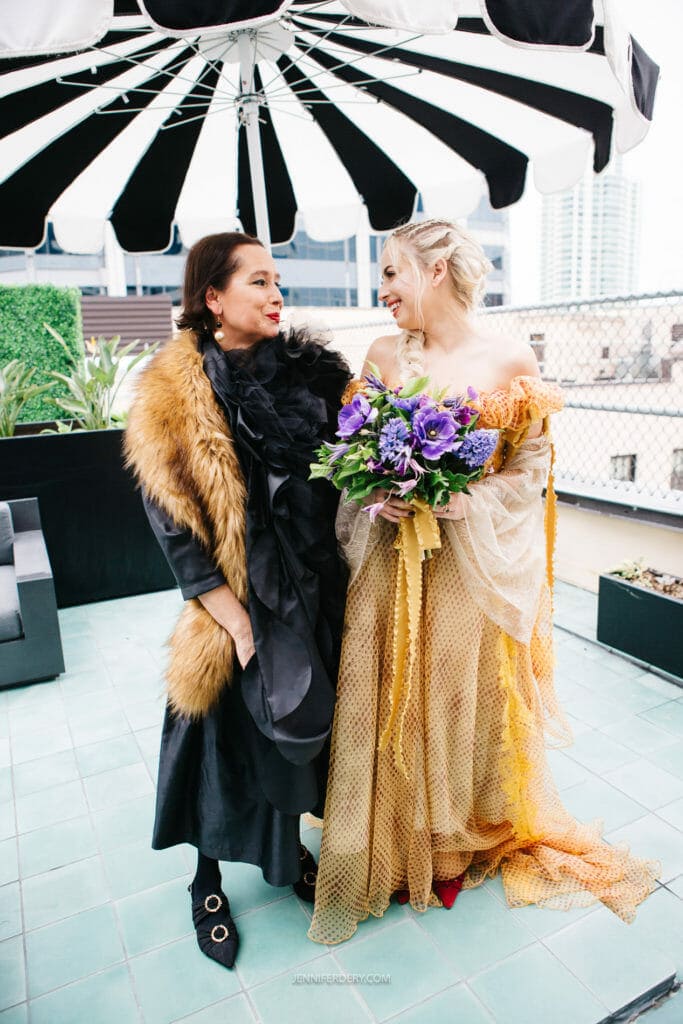Two women standing outdoors on a tiled floor, under a large black and white striped umbrella. The woman on the left wears a black dress with a brown fur stole, while the woman on the right wears a golden, off-the-shoulder gown and holds a bouquet of purple flowers. Both smile at each other.