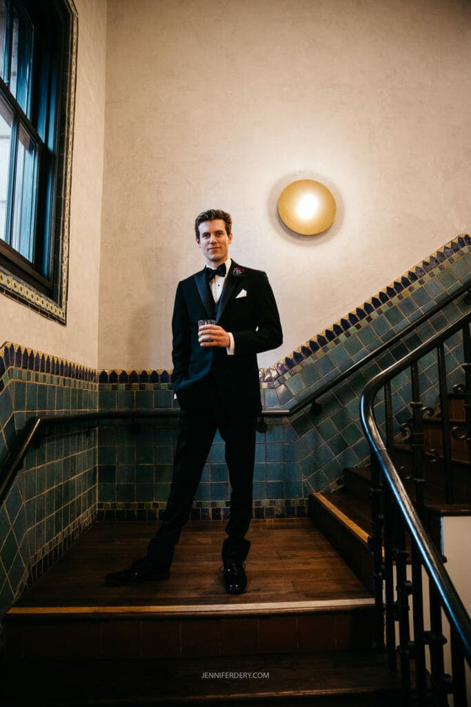 A groom in a black tuxedo stands confidently on a stairway holding a drink. He is positioned in front of a wall adorned with tiles and a circular light fixture. The decor has an elegant, classic feel.