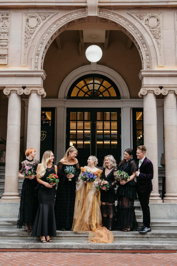 A bride in a golden gown stands on steps outside a building with a group of bridesmaids and groomsmen. The bridesmaids are dressed in black and hold colorful bouquets while the groomsmen, wearing black suits, hold their hands behind their backs.