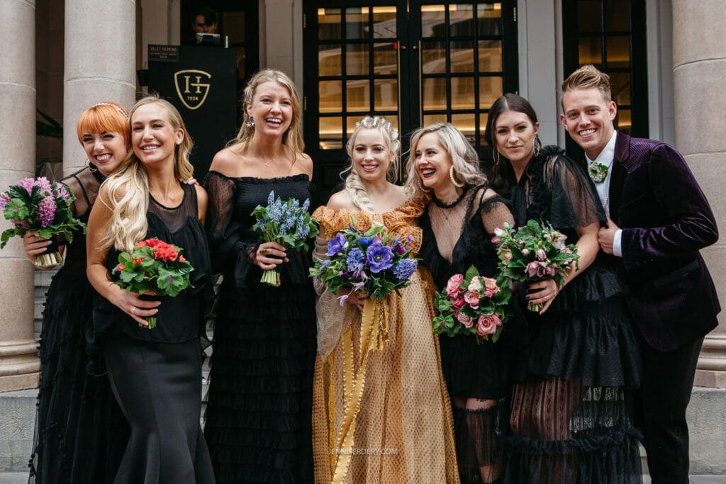 A bridal party, including six women and one man, stand smiling on stone steps wearing formal attire. The woman in the center wears a gold dress and holds a bouquet of purple flowers. The others are dressed in black and hold bouquets of red and pink flowers.