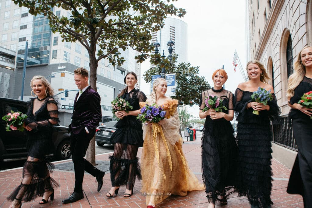 A joyful group of individuals walks down a city sidewalk. The group, mostly women, wear stylish black dresses and carry floral bouquets. One person stands out in a gold dress. Background features tall buildings, trees, and parked cars.