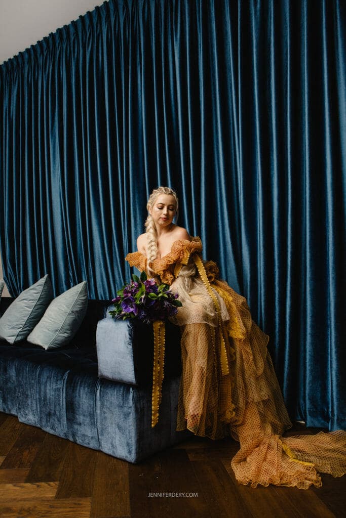 A bride in an elaborate, ruffled gown with a long train sits on a blue velvet couch. She holds a bouquet of purple flowers and is positioned against a backdrop of heavy, dark blue curtains. Several light blue pillows are visible beside her on the couch.