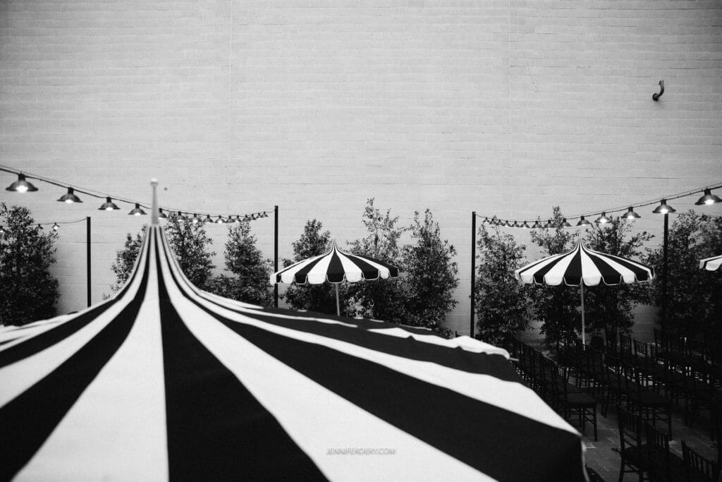Monochromatic outdoor space featuring black-and-white striped umbrellas, string lights, and rows of empty chairs in front of a white brick wall. The setup suggests a venue prepared for a gathering or event.