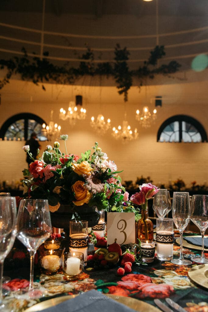A beautifully set dining table adorned with an elegant floral centerpiece displaying a variety of colorful flowers. The table is decorated with glassware, candles, and a table number card with the number 3. Hanging chandeliers provide soft lighting, enhancing the ambiance.