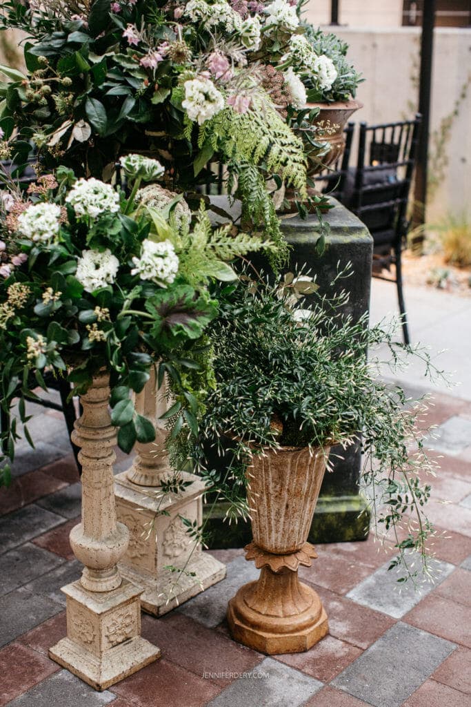 Tall floral arrangements in ornate vases, featuring lush greenery and white flowers, stand on a tiled patio. A weathered stone pedestal supports one arrangement, while another planter displays cascading green foliage. Black chairs are visible in the background.