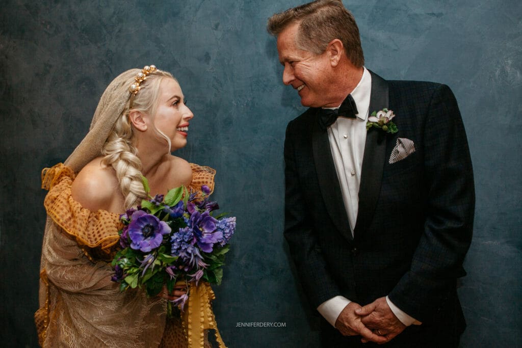 A bride and her dad, both dressed formally, smile at each other. The bride holds a bouquet of purple flowers and wears a cream-colored dress with a lace shawl. The man, in a black tuxedo with a checkered pocket square, appears to be her father. They stand against a dark blue textured background.