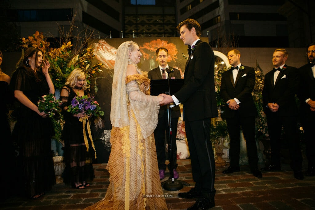 A couple stands facing each other at an outdoor nighttime wedding ceremony. The bride is wearing a flowing gown and veil, holding the groom's hands. The groom is in a black tuxedo. The officiant stands behind a lectern. Bridesmaids and groomsmen are beside them.