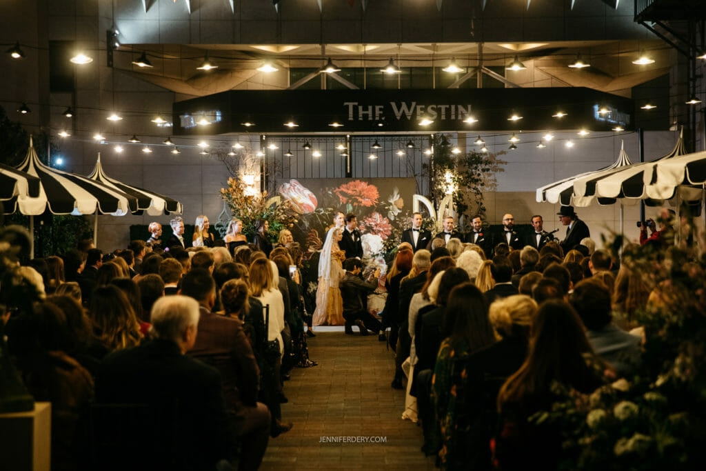 A wedding ceremony is taking place at night, and outside under the stars. Guests are seated on both sides, facing the wedding party at the front. The space is decorated with flowers and plants, and overhead lights create a warm ambiance.