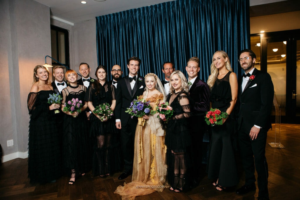A group photo of a wedding party indoors. The bride, center, in a white dress holds a bouquet, surrounded by the groom and attendants in formal attire, some holding bouquets of flowers. The background features dark blue curtains and a wooden floor.
