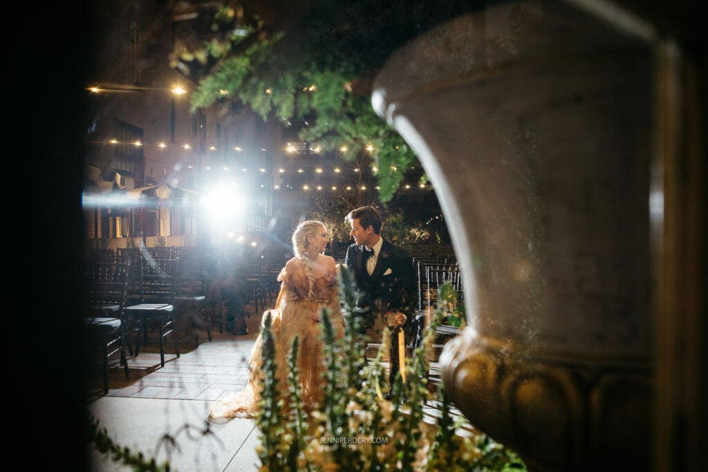 A couple dressed elegantly is sitting on a bench in an outdoor setting, illuminated by warm string lights. The surroundings have a romantic ambiance, with empty chairs in the background and plants in the foreground. The woman is in a light-colored dress, and the man is in a suit.