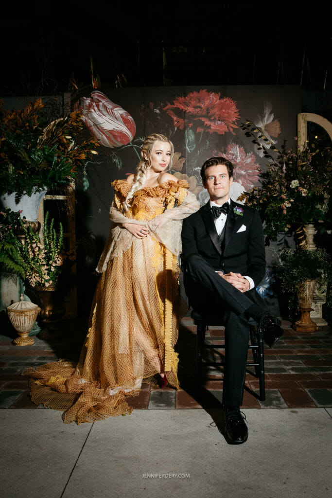 A woman in a golden dress adorned with flowers and a man in a black tuxedo pose for a formal photo against a dark floral backdrop. The woman stands while the man sits on a stool. The setting is elegantly decorated with large plants and floral arrangements.