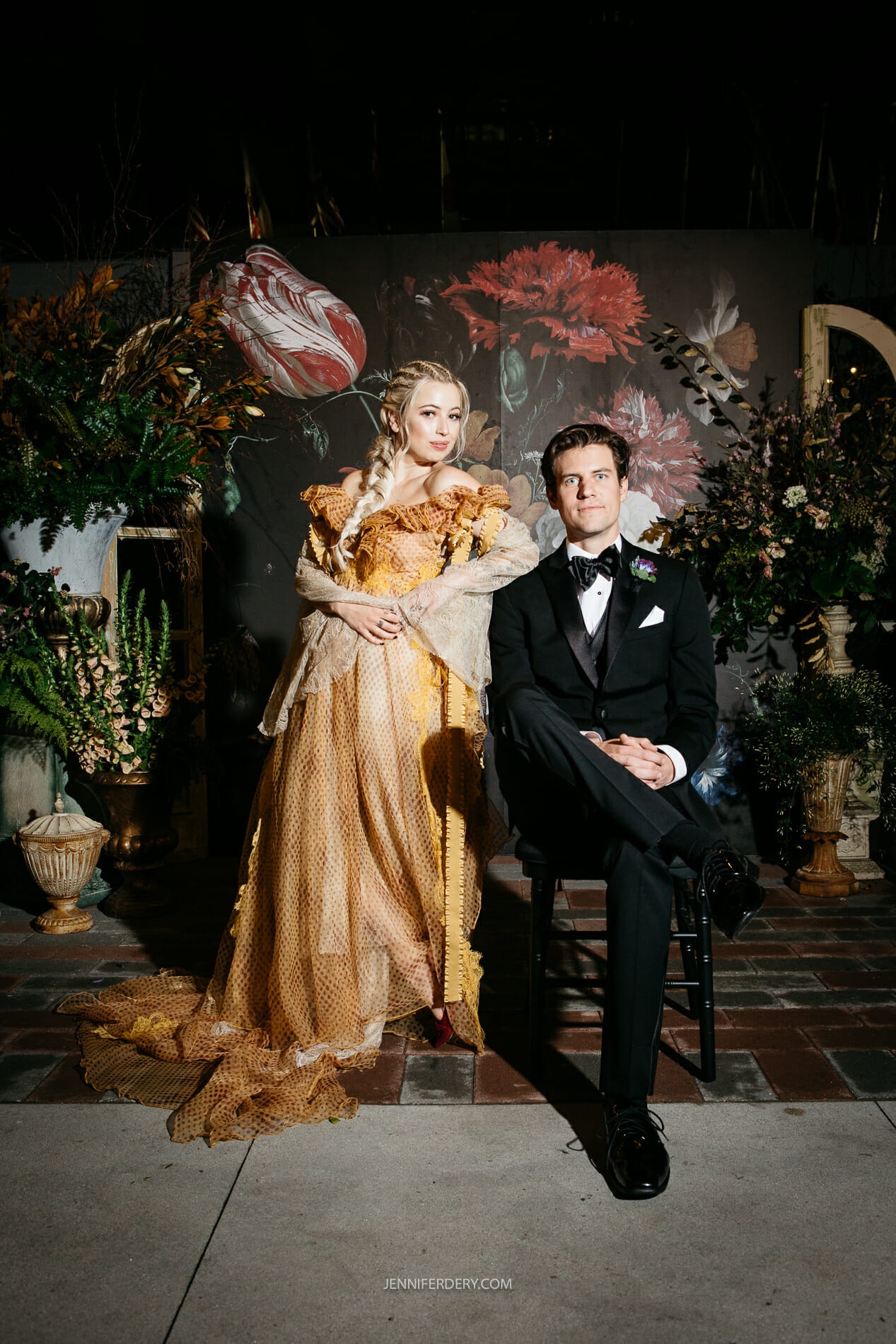 A woman in a golden dress adorned with flowers and a man in a black tuxedo pose for a formal photo against a dark floral backdrop. The woman stands while the man sits on a stool. The setting is elegantly decorated with large plants and floral arrangements.