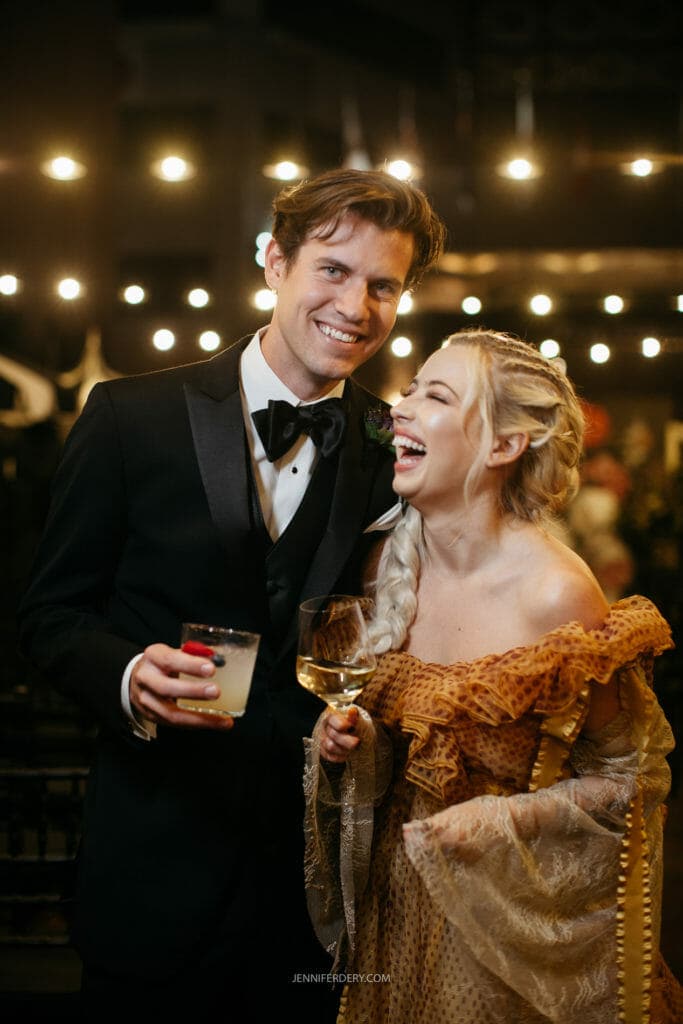 A man and woman are dressed elegantly, smiling and laughing together at an event. The man is wearing a black tuxedo and holding a drink, while the woman is in a gold off-the-shoulder dress, also holding a glass. String lights are visible in the background.