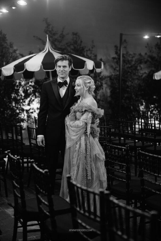 A black and white photo features a couple standing closely together in a dimly lit, elegant outdoor setting. The man wears a black tuxedo, and the woman is dressed in a unique, ruffled gown. They are surrounded by rows of empty chairs and shaded by large, decorative umbrellas.