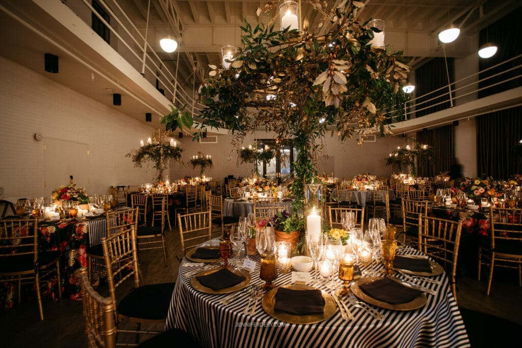 A beautifully decorated venue set up for a wedding reception. The tables are adorned with striped tablecloths, elegant gold chairs, and a variety of flowers and candles. Hanging decorations and lights add to the warm, festive ambiance.