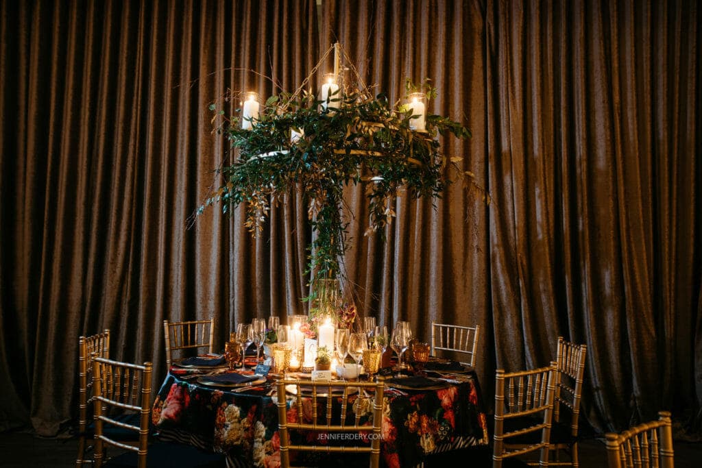 A round dining table elegantly set with floral tablecloth, candles, and glassware. Above, an elaborate chandelier adorned with greenery and illuminated candles hangs. The background features long, luxurious brown drapes. Golden chairs surround the table.