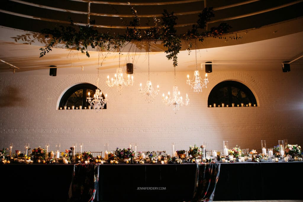 A beautifully decorated event space features a long banquet table adorned with candles, floral arrangements, and elegant tableware. Chandeliers and fairy lights hang from the ceiling, and a white brick wall with arched windows forms the backdrop.