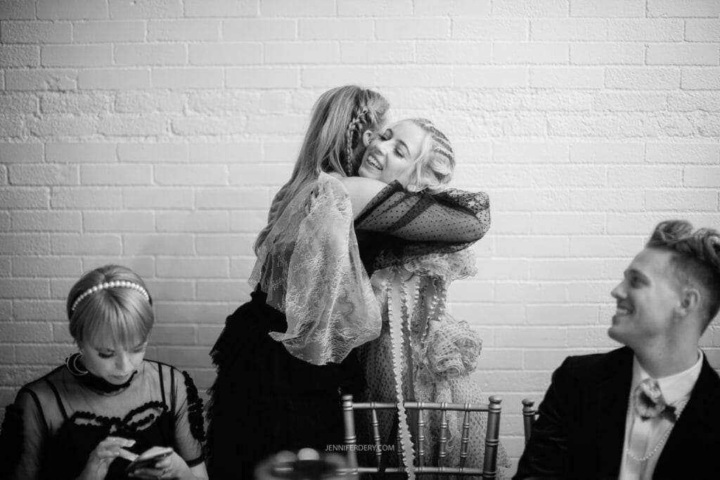 Black-and-white photo of four people dressed in formal, possibly vintage attire. Two women in the center share a joyful embrace. To their left, a woman sits and looks down at something in her hands. To their right, a man sits, smiling.