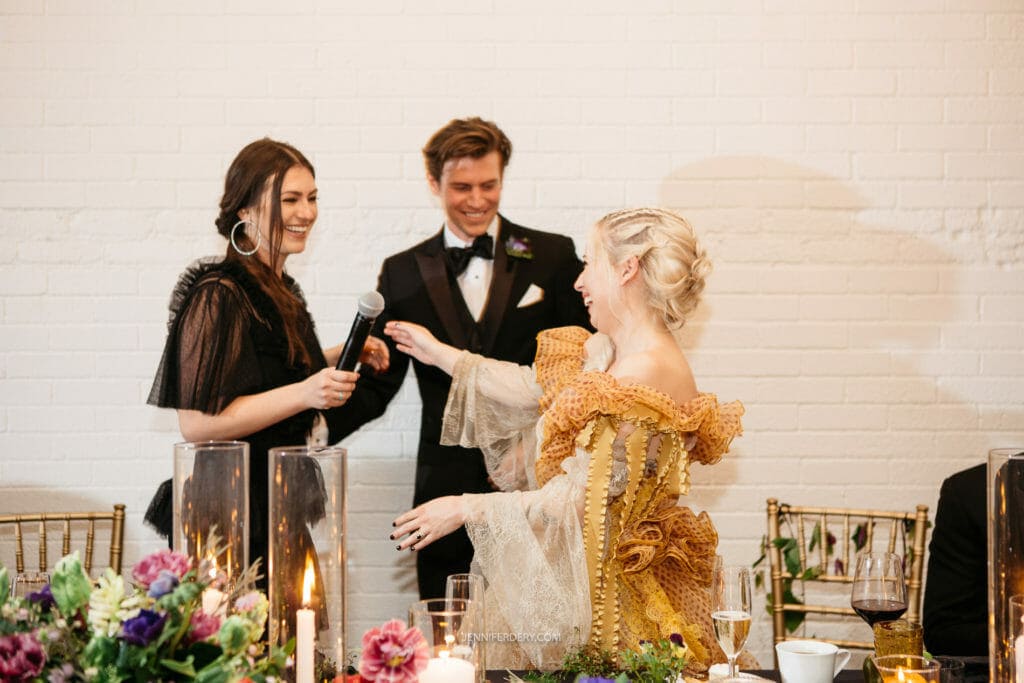 A woman in a black dress stands holding a microphone, speaking to a smiling couple dressed in formal attire. The man wears a black tuxedo, while the woman dons an ornate, golden off-the-shoulder dress. They stand in front of a white brick wall, surrounded by a decorated table.