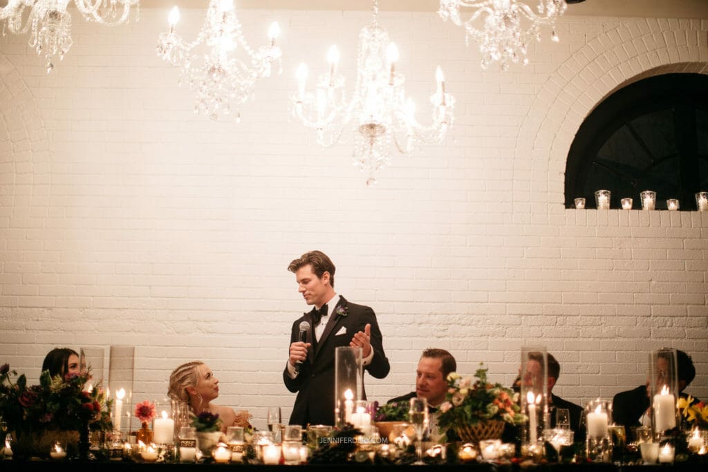 A groom in a tux stands and speaks into a microphone at a formal event, possibly a wedding, with elegantly decorated tables and chandeliers above. Seated guests, dressed formally, listen and look on at the speaker. Lit candles and flower arrangements adorn the tables.