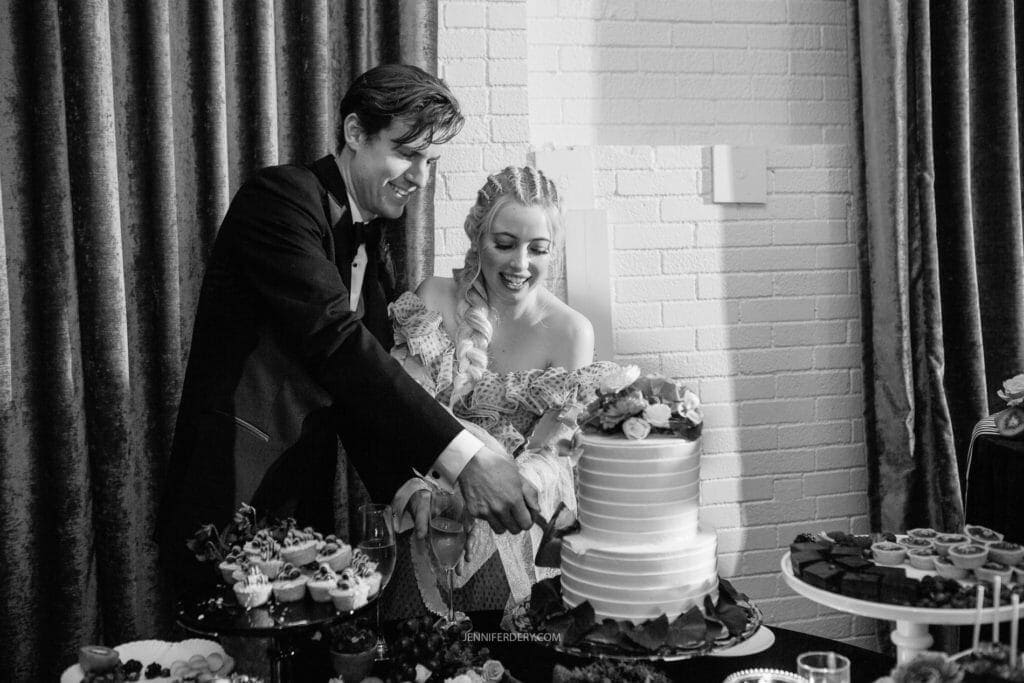 A couple dressed in formal attire smiles as they cut a tiered wedding cake together. The man wears a black tuxedo and the woman is in a wedding dress with a floral bouquet in her hand. Various desserts are displayed on the table beside the cake. The background features a brick wall and curtains.