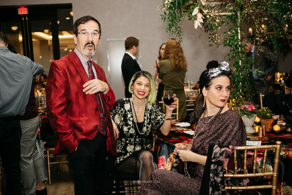 Three people are dressed in formal attire, attending a festive indoor event. A man in a red jacket and glasses stands with two women. One woman, with short green hair, is smiling while holding a wine glass, and the other woman is seated, looking to the side.