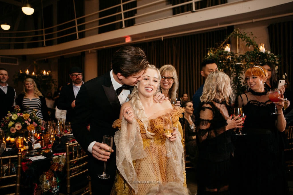 A couple dressed formally shares a joyful moment at a celebration, with the man kissing the woman's forehead. The woman holds a glass of champagne and smiles. They are surrounded by smiling guests who are also holding drinks in a warmly lit venue decorated with flowers.