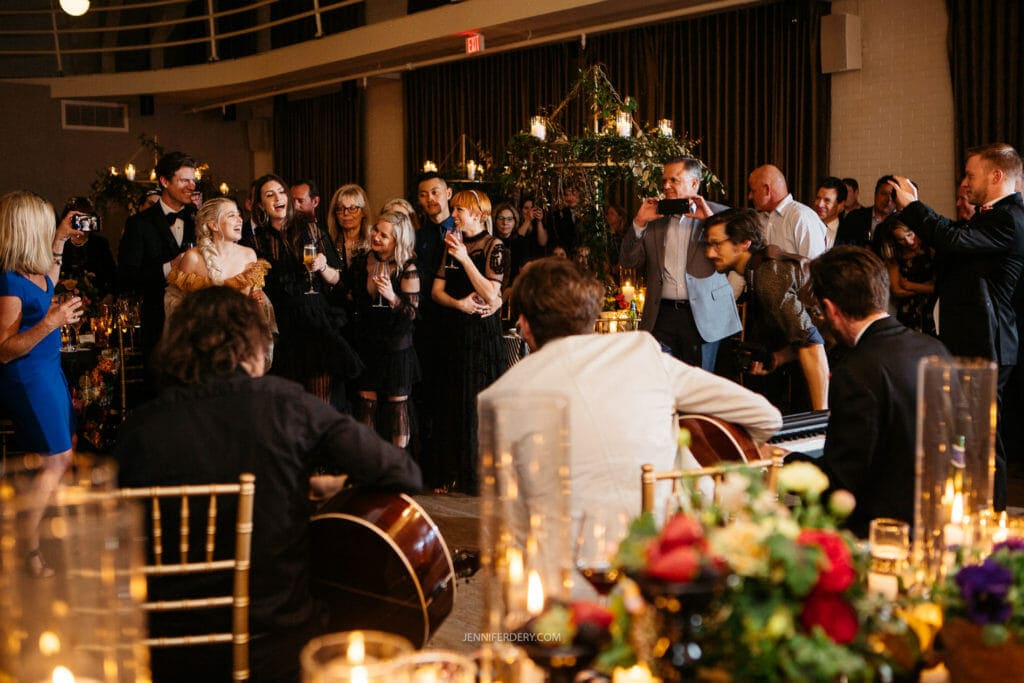 A group of people dressed in formal attire are gathered in a warmly lit room, enjoying a musical performance. Flowers and candles adorn tables. Some guests are smiling, taking photos, and holding drinks while musicians play guitars in the foreground.
