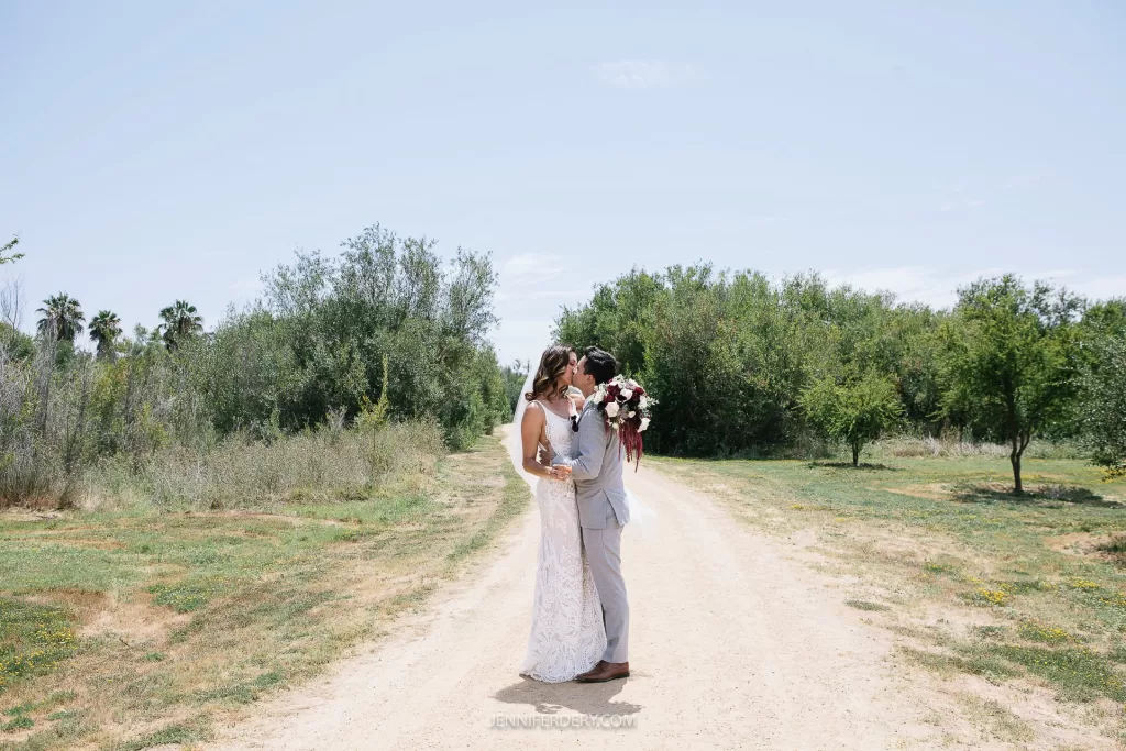 A couple dressed in wedding attire kisses on a dirt path surrounded by greenery in rancho guajome park. The bride holds a bouquet of flowers, and they are embraced under a clear sky.