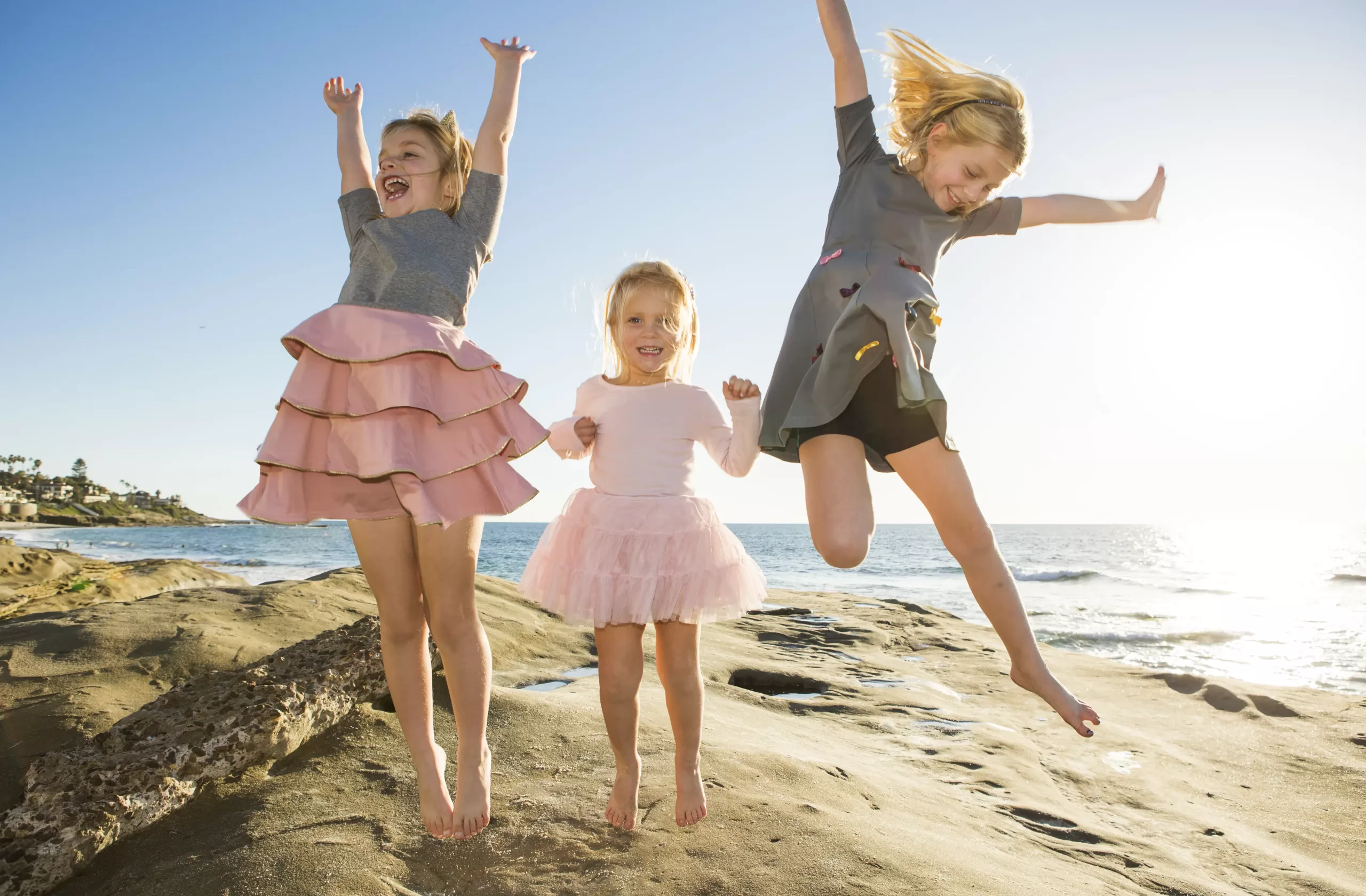 Three young girls are happily jumping on a sunlit beach, dressed in playful skirts and dresses. The ocean waves and a blue sky create a picturesque backdrop, with the sunlight casting a warm glow over the scene. Everyone appears to be enjoying the moment.