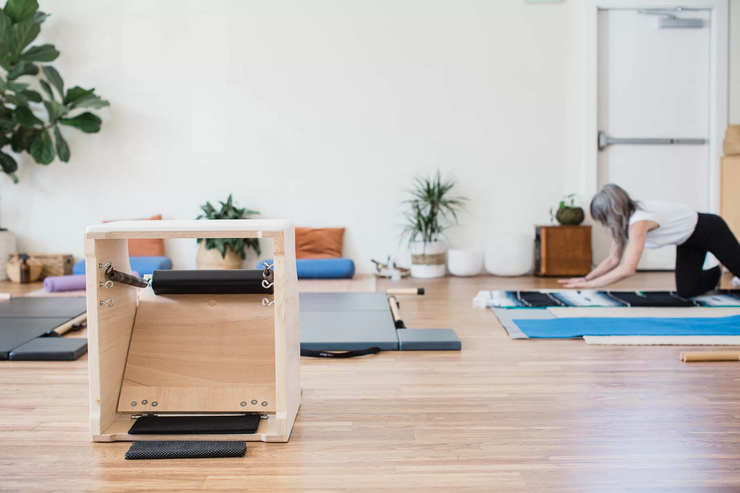 A serene Pilates studio captured by a San Diego branding photographer showcases a wooden exercise apparatus in the foreground and a person with gray hair in athletic wear stretching on a mat in the background. The studio features wooden floors, minimalistic decor, and potted plants.