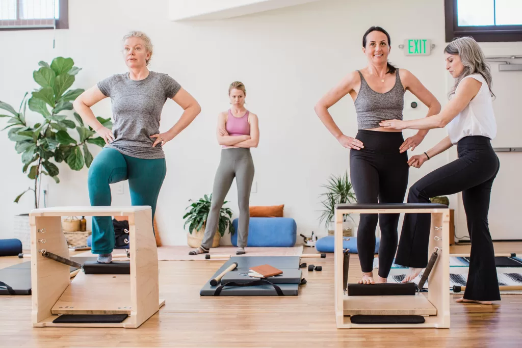 In a well-lit studio, captured by a San Diego branding photographer, women participate in a vibrant fitness class. Two step on exercise equipment with guidance from an instructor, while others stand in the background. Various gym accessories and potted plants enhance the energetic setting.