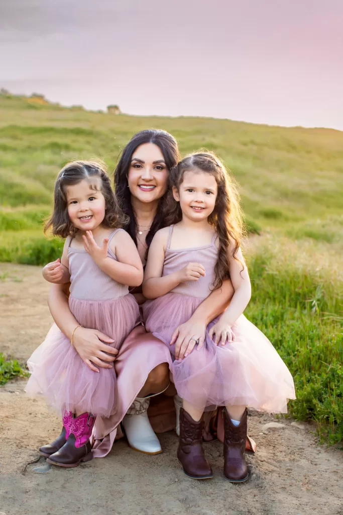 mother's day session showing A woman kneels on a dirt path in a grassy field at sunset, smiling and holding two young girls wearing matching light purple dresses and cowboy boots. The girls have curly hair and are both smiling, with the woman positioned directly behind them.