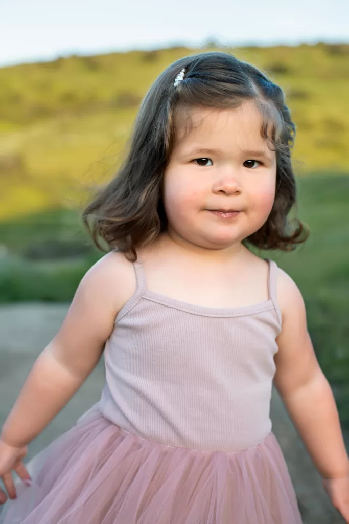 mother's day session showing A young girl with wavy brown hair stands outdoors on a sunny day. She is wearing a light pink tank top and a matching pink tulle skirt. The background features a green, grassy landscape. She has a slight smile on her face.