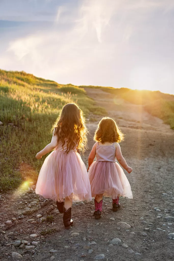 mother's day session showing two young girls wearing pale pink dresses walk hand in hand up a dirt path towards the sunset. The path is flanked by grassy hills, and the sun casts a warm glow, illuminating their hair and surroundings with a soft, golden light.