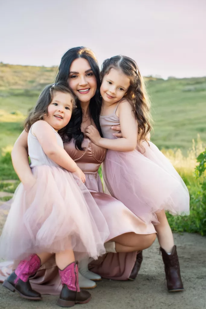 mother's day session showing a smiling woman crouches outdoors with two young girls, all wearing light-colored dresses. The woman and children embrace each other affectionately. The background features a grassy hillside with a serene, early evening sky. The girls wear cowboy boots.