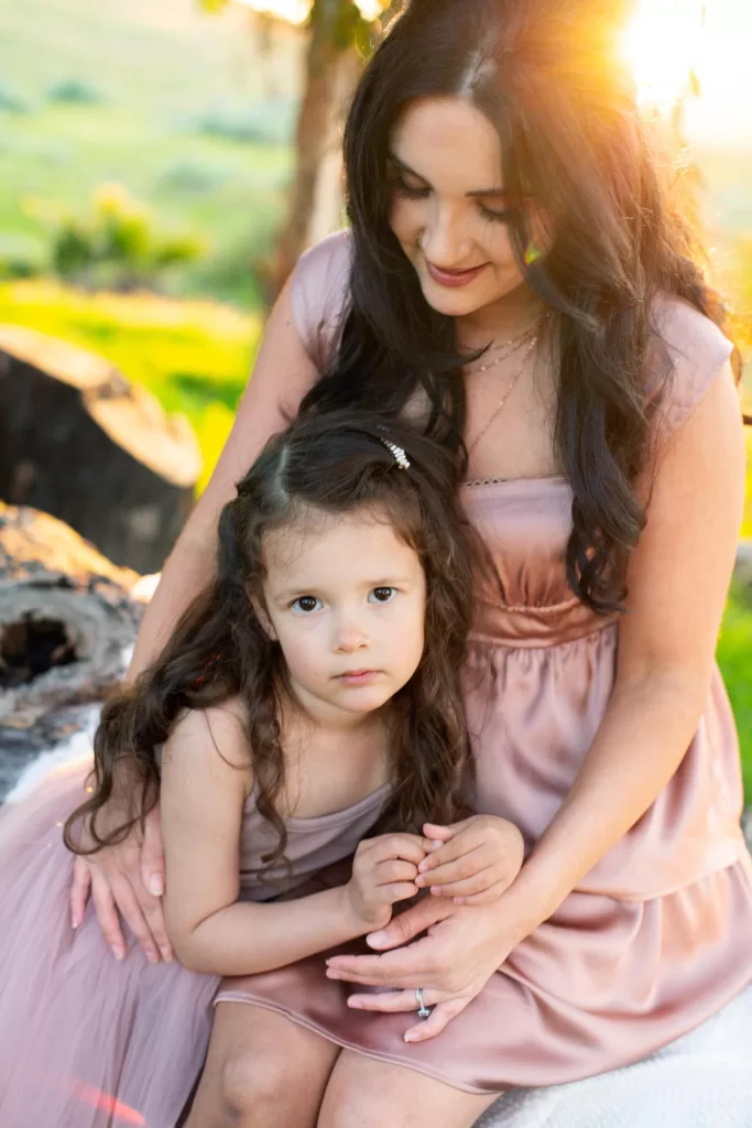 mother's day session showing A woman with long dark hair, wearing a pink dress, sits with a young girl with similar hair, also in a pink dress. They are outdoors during sunset, with light filtering through trees in the background. The woman holds the girl's hands gently, and both appear calm.