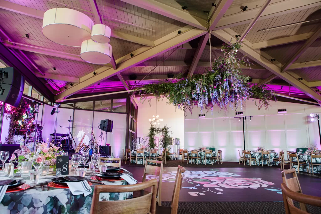photo of the ballroom at the university club in palo alto set up for a bat mitzvah reception. includes a large flower installation on the ceiling, a stage for a band and a black dance floor embellished with a giant floral design