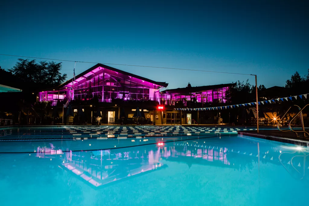 photo of the university club at dusk. building is lit inside by purple lights and is reflected in the large blue swimming pool in the foreground.