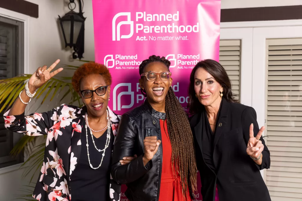 Three women stand in front of a Planned Parenthood banner, smiling and gesturing with peace signs. The woman on the left wears a floral blazer, the woman in the center has braids and glasses, and the woman on the right is in a black blazer.