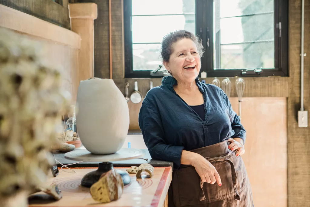 A woman in a blue shirt and brown apron stands smiling in a workshop. She is near a table with pottery tools and a large clay vase. The workshop has brown walls and windows letting in natural light. She appears relaxed and happy.