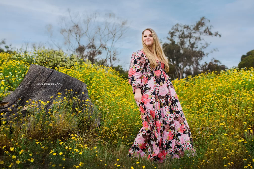 A woman stands in a field of yellow wildflowers wearing a long, floral dress. She has long blonde hair and looks off into the distance. There is a tree stump to her left and a background of trees and a blue sky.
