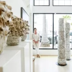 A person stands in a bright, modern gallery filled with unique, textured ceramic sculptures. Large windows behind them let in natural light, illuminating the white walls and wooden floor. Two tall ceramic columns are displayed prominently in the foreground.