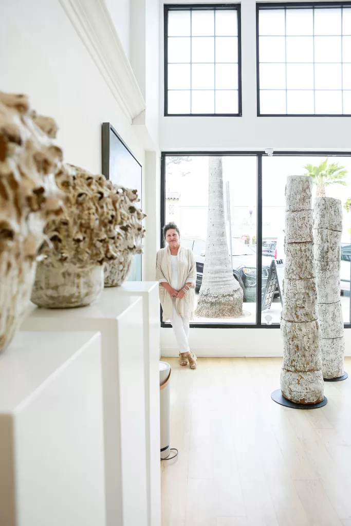A person stands in a bright, modern gallery filled with unique, textured ceramic sculptures. Large windows behind them let in natural light, illuminating the white walls and wooden floor. Two tall ceramic columns are displayed prominently in the foreground.