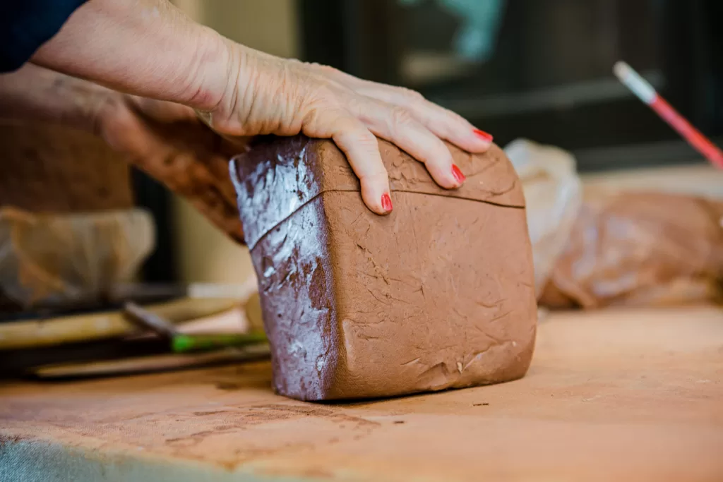 Close-up of a person's hand with red painted nails shaping a large block of clay on a table. Various pottery tools and another block of clay are visible in the background. The setting appears to be a pottery workshop.
